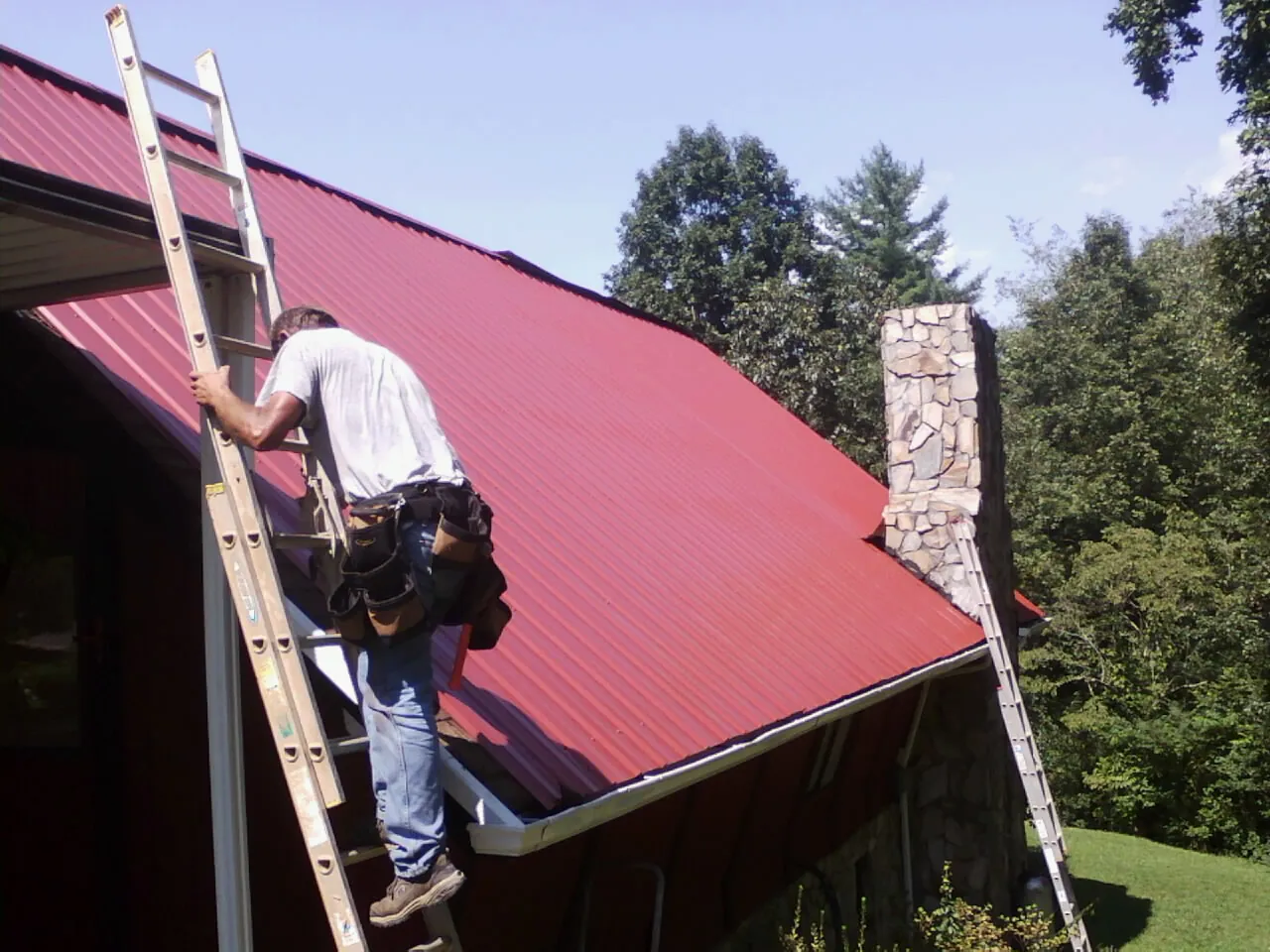 Roofer on a ladder beside a red metal roof and stone chimney — Soffit & Fascia Repair by Roofing Brothers in Highland Mills